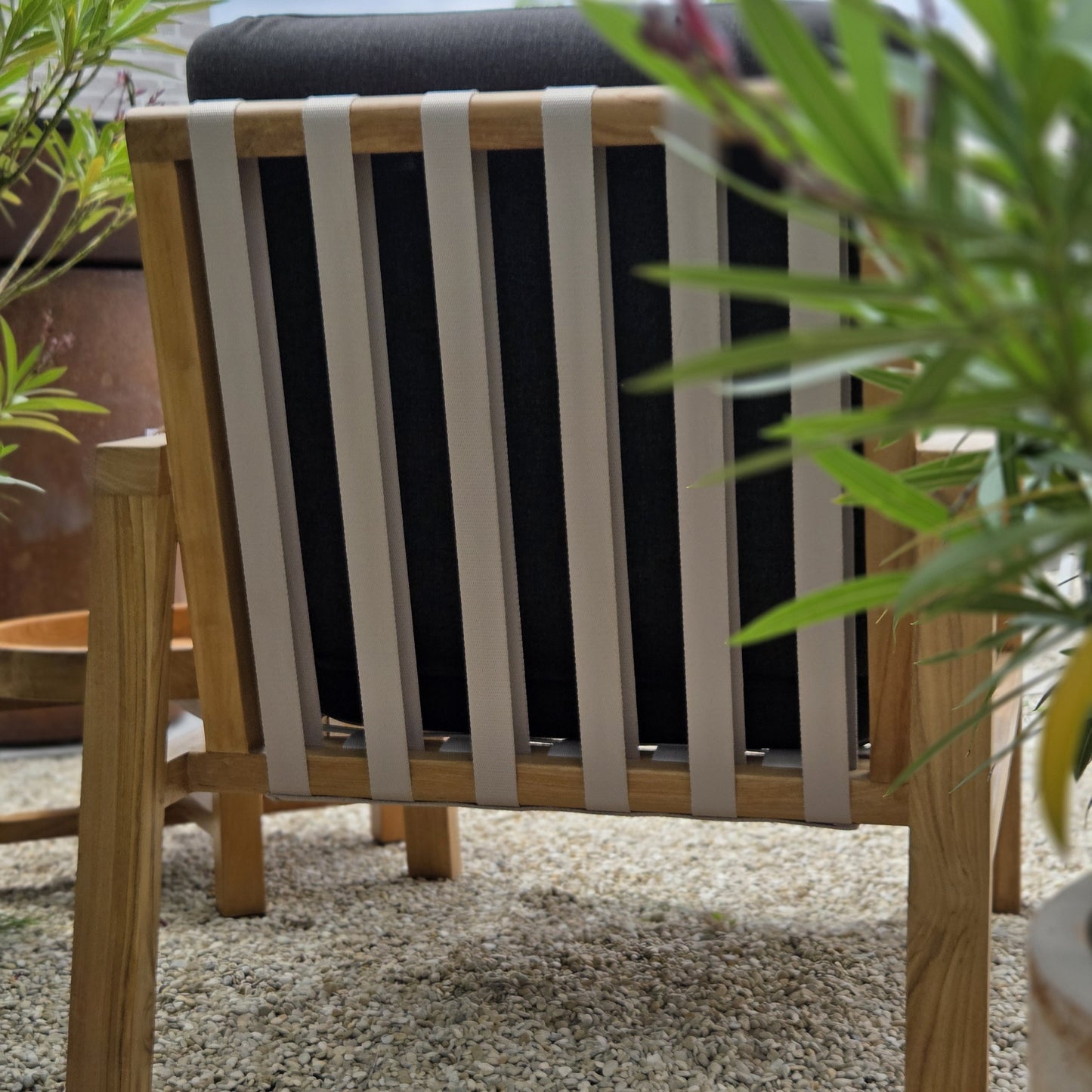 Rear view of a teak outdoor lounge chair featuring woven support straps and black cushions in a garden setting