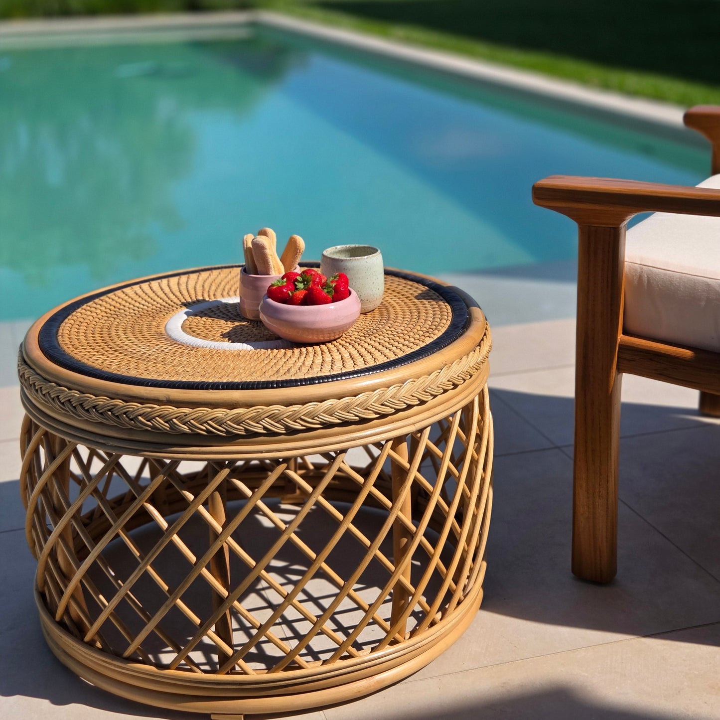 Mesa auxiliar tejida de ratán, decorada con fresas frescas, junto a un sillón lounge de teca de grado A con vistas a una piscina mediterránea.
