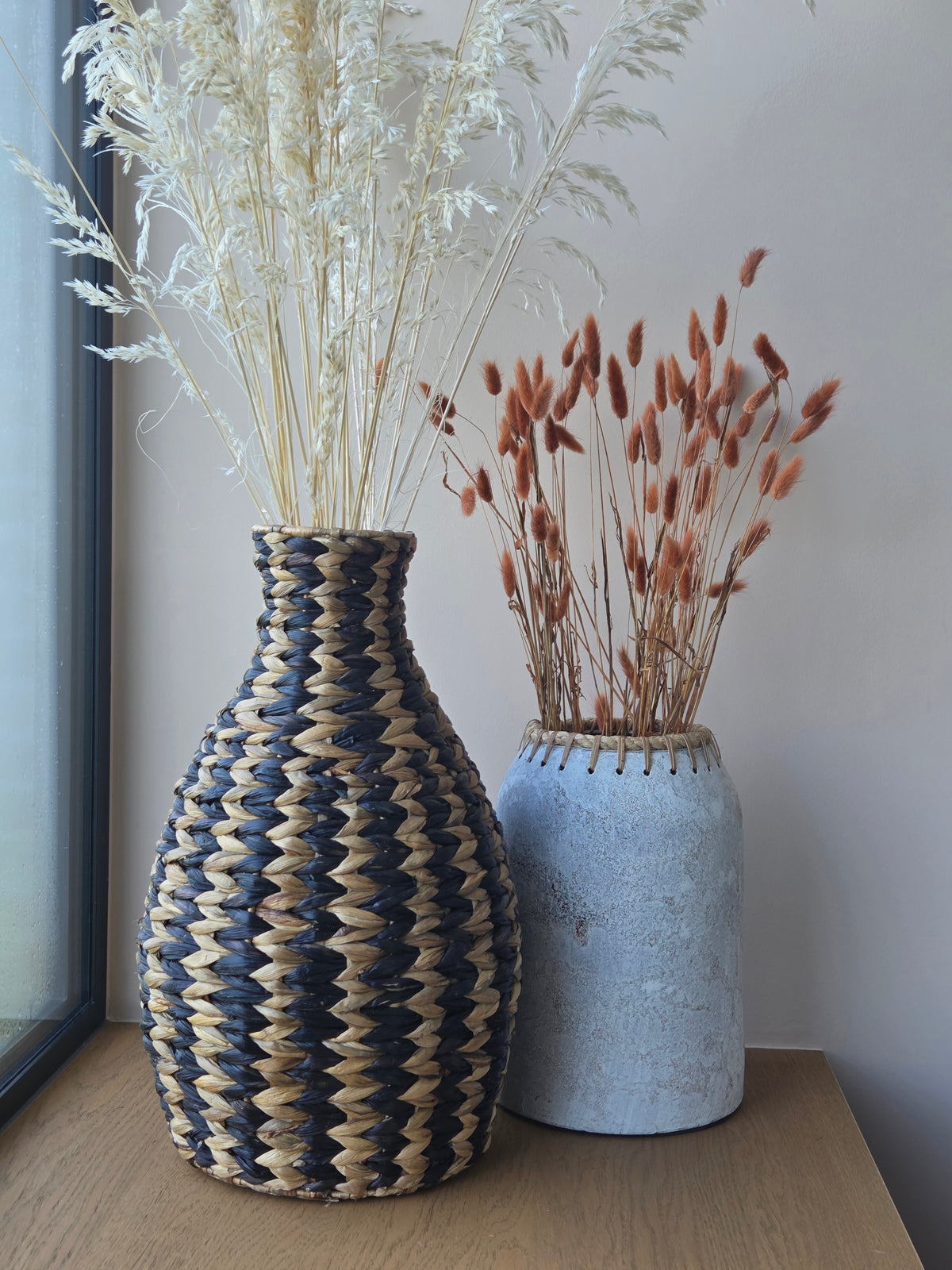 Two decorative vases styled with dried flowers: a black and natural woven vase with a geometric pattern and a rustic grey vase with textured finish, displayed on a wooden surface.