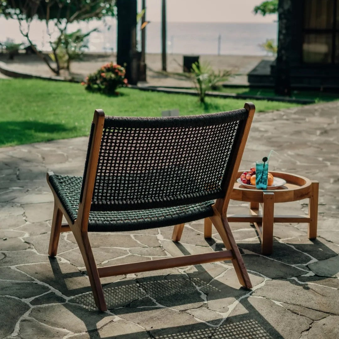 Siga-Siga outdoor lounge chair with a handcrafted teak frame and black synthetic rattan weave, styled on a sunlit patio near the ocean.