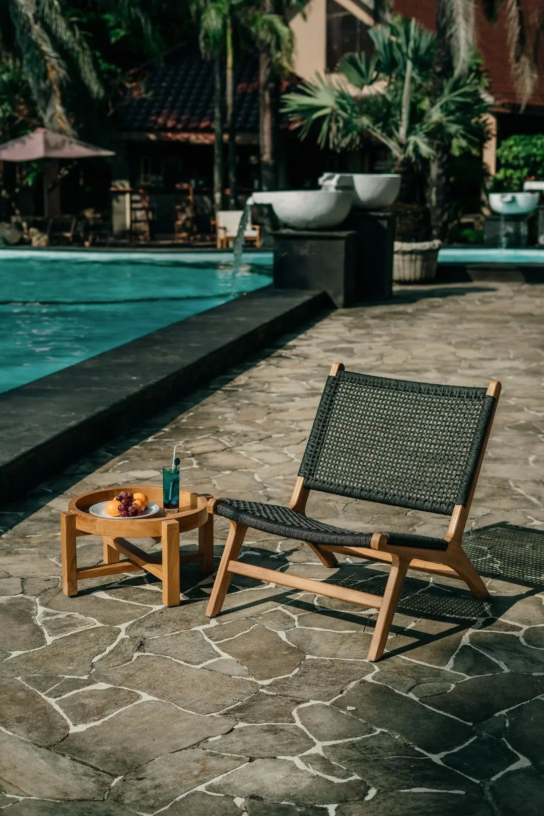 Siga-Siga outdoor lounge chair with a handcrafted teak frame and black synthetic rattan weave, styled beside a swimming pool with a matching teak side table.
