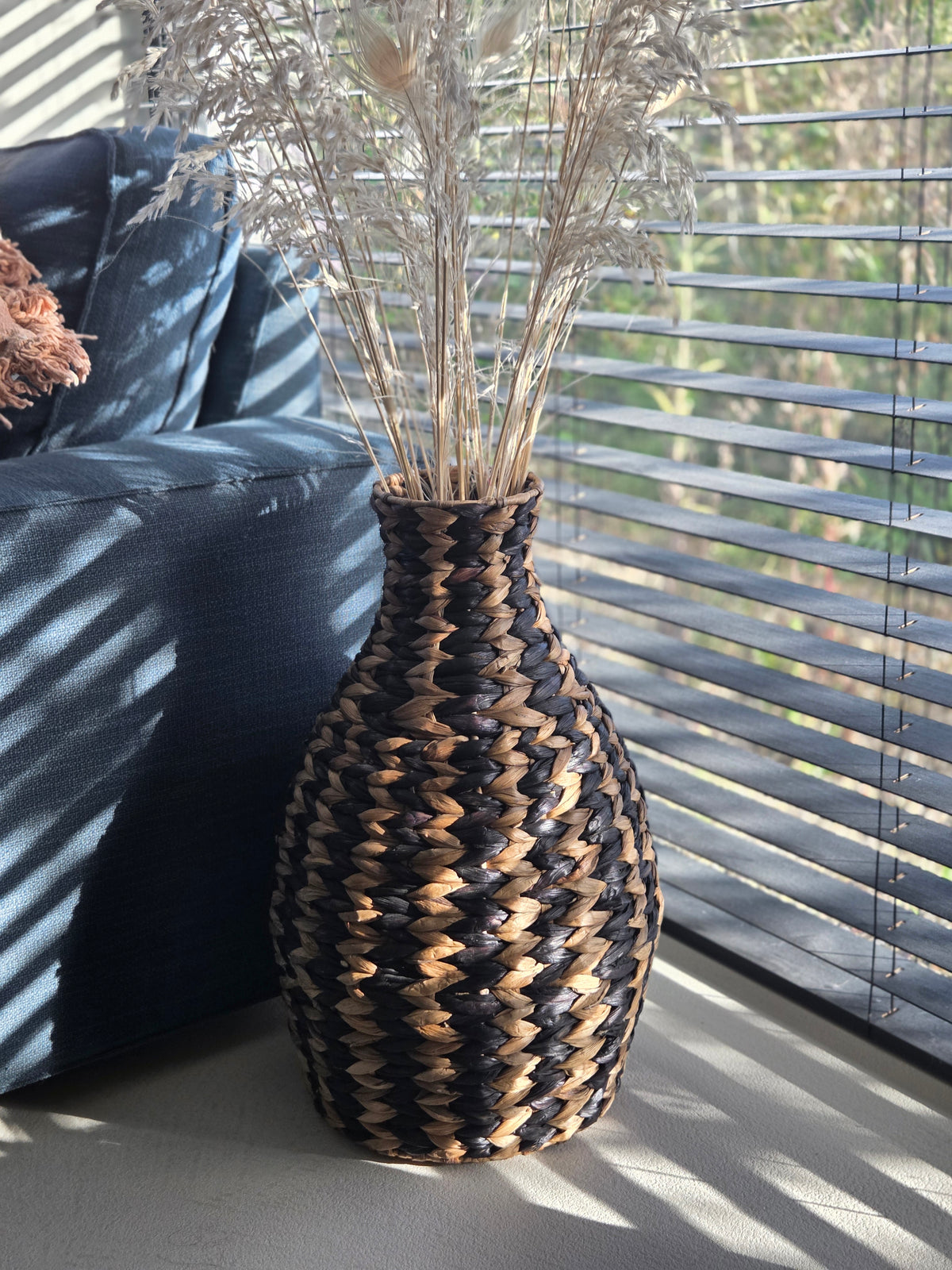 Jarrón decorativo de suelo de fibras tejidas en negro y color natural, con un diseño geométrico, decorado con hierbas secas junto a un sofá oscuro en una sala de estar iluminada por el sol.