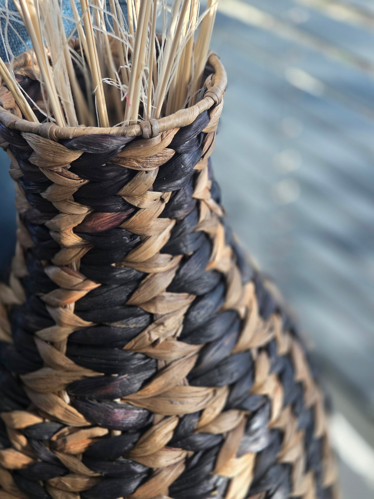 Close-up of the narrow neck and braided rim of a black and natural woven vase, highlighting the contrasting fibres and intricate geometric weave.