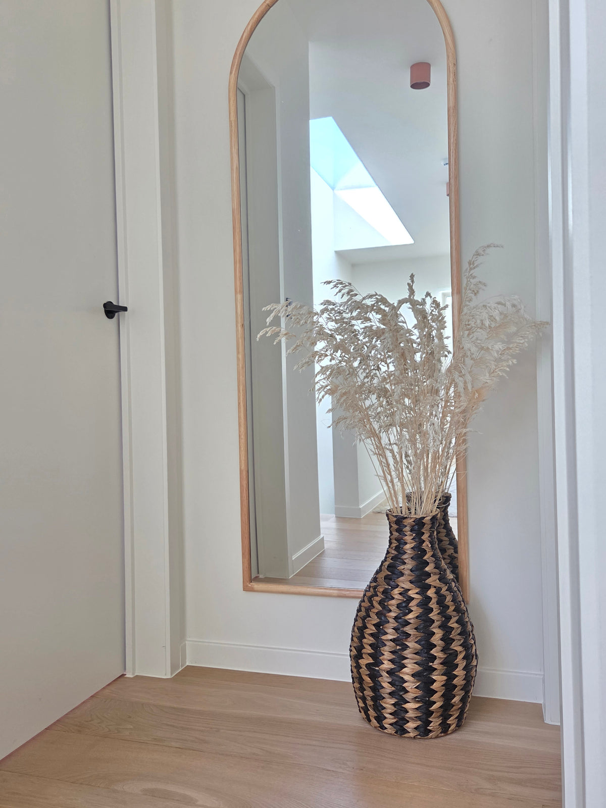 Zahara black and natural woven floor vase filled with dried grasses, displayed in a minimalist hallway beneath a tall arched wooden mirror.