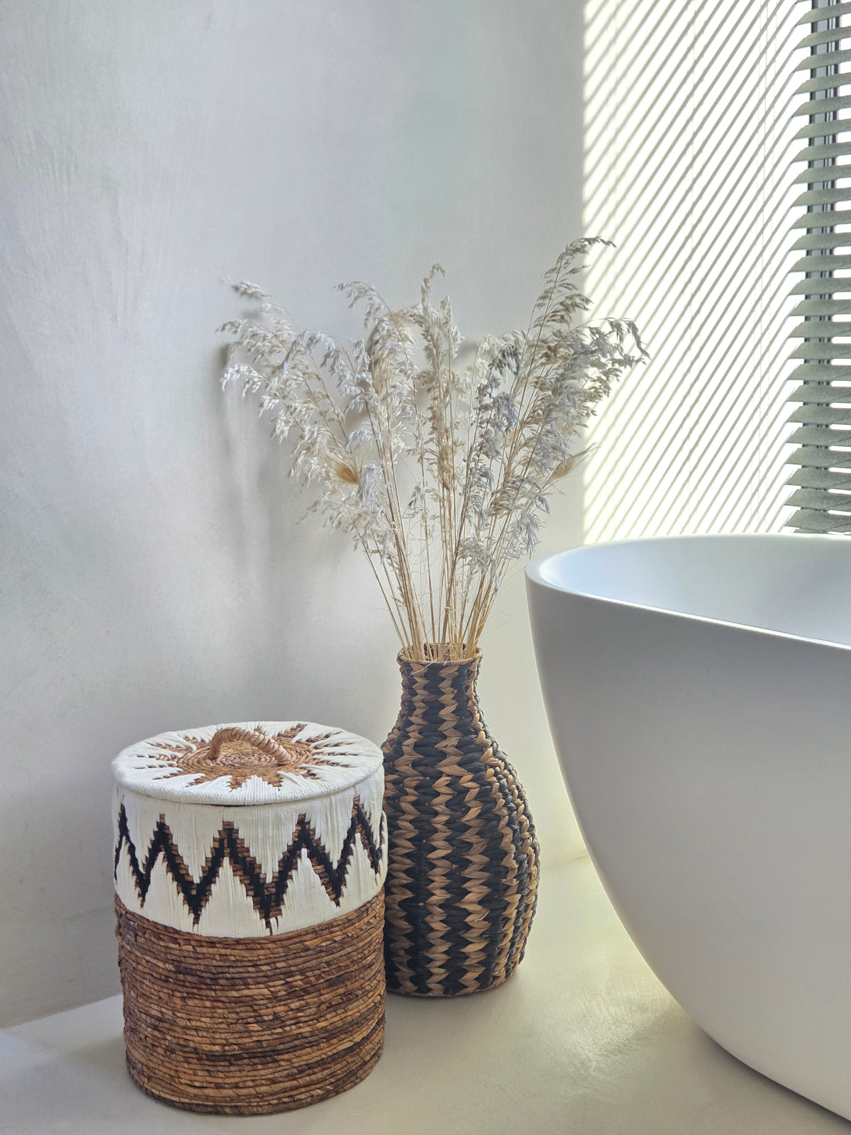 Zahara black and natural woven decorative vase styled with dried grasses beside a patterned storage basket and a white freestanding bathtub in a sunlit bathroom