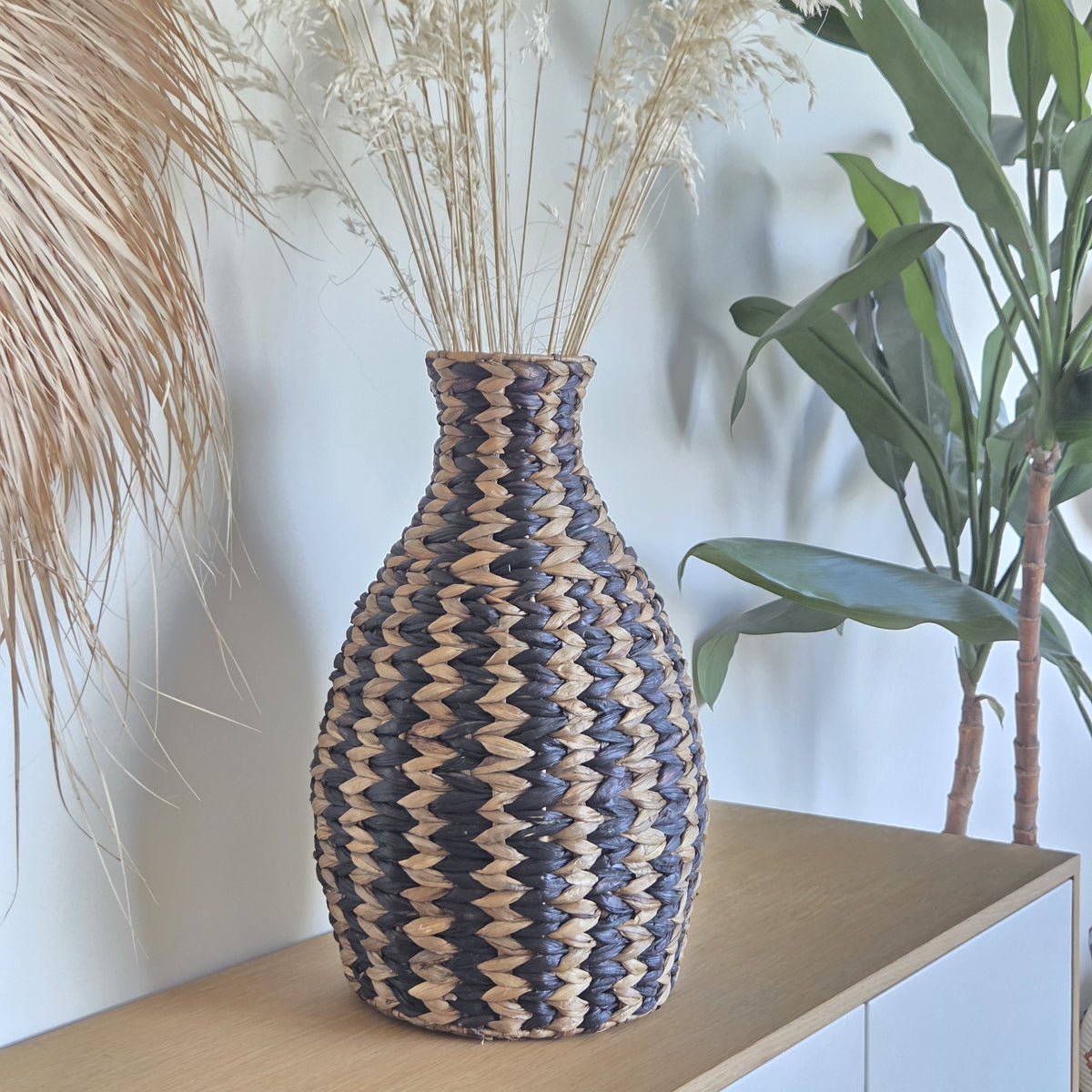 Zahara decorative woven vase featuring a black and natural zigzag pattern, styled with dried grasses on a wooden cabinet in a bright, contemporary interior