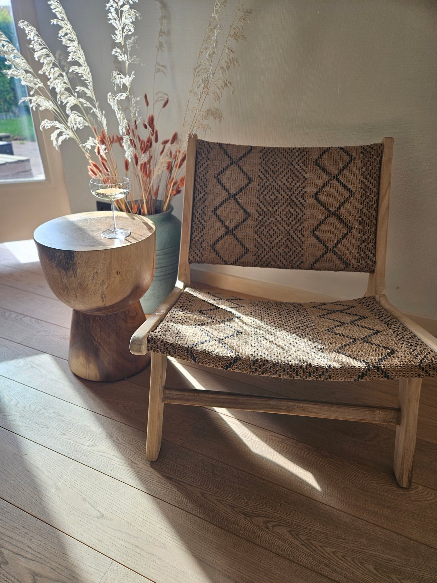 Funchal lounge chair with a handcrafted teak wood frame and woven textile seat, styled with a wooden side table and dried grasses in a warm organic modern interior.