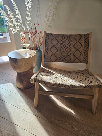 Funchal lounge chair with a handcrafted teak wood frame and woven textile seat, styled with a wooden side table and dried grasses in a warm organic modern interior.