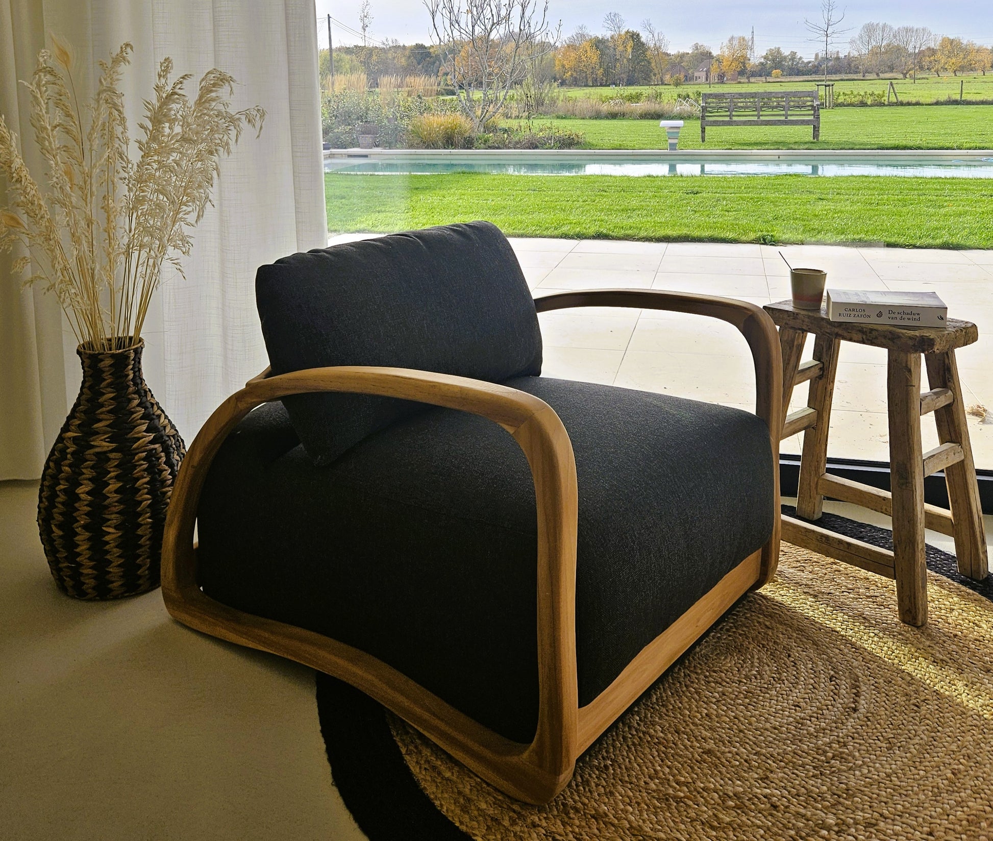Modern teak lounge chair with dark upholstery in a bright natural interior, styled with a woven rug, handcrafted vase and garden views.