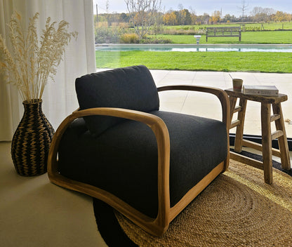 Modern teak lounge chair with dark upholstery in a bright natural interior, styled with a woven rug, handcrafted vase and garden views.
