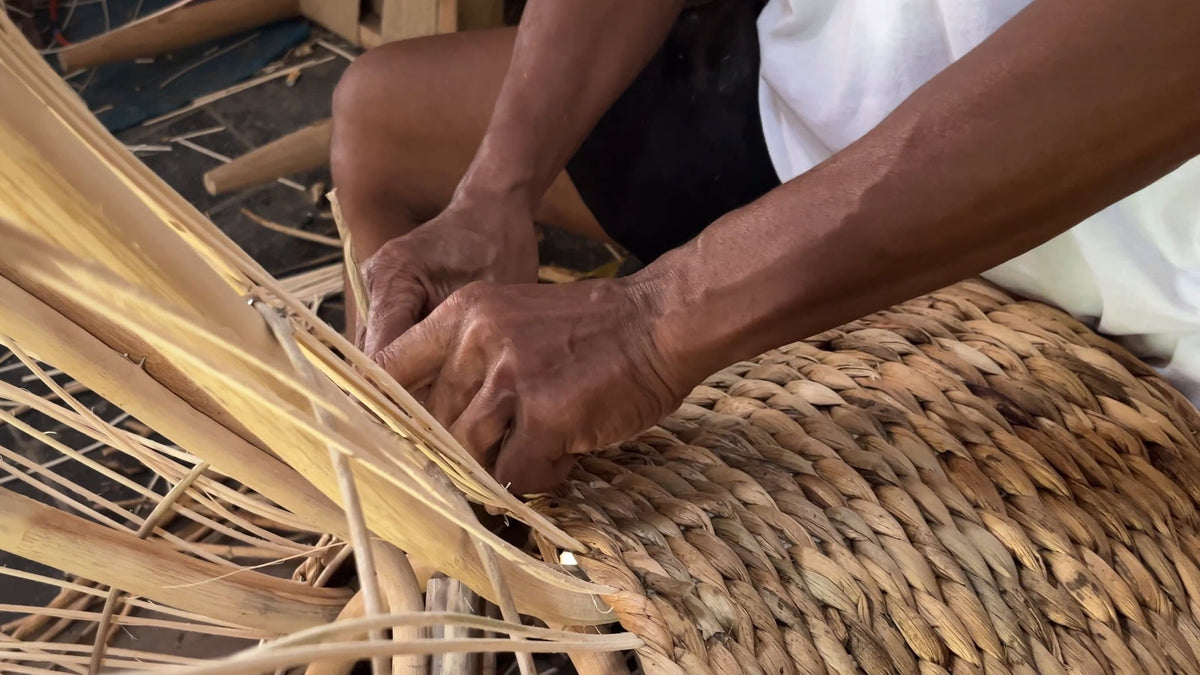 Primer plano de una artesana indonesia tejiendo a mano fibras secas de jacinto de agua para crear una mesa de centro, que muestra la artesanía tradicional y las texturas naturales del tejido.