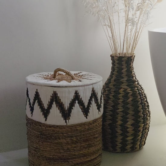 Samuel White handwoven abaca storage basket featuring a distinctive black and white zigzag pattern, a fitted lid and a decorative handle, styled beside the Zahara woven vase in a bright, natural bathroom interior.