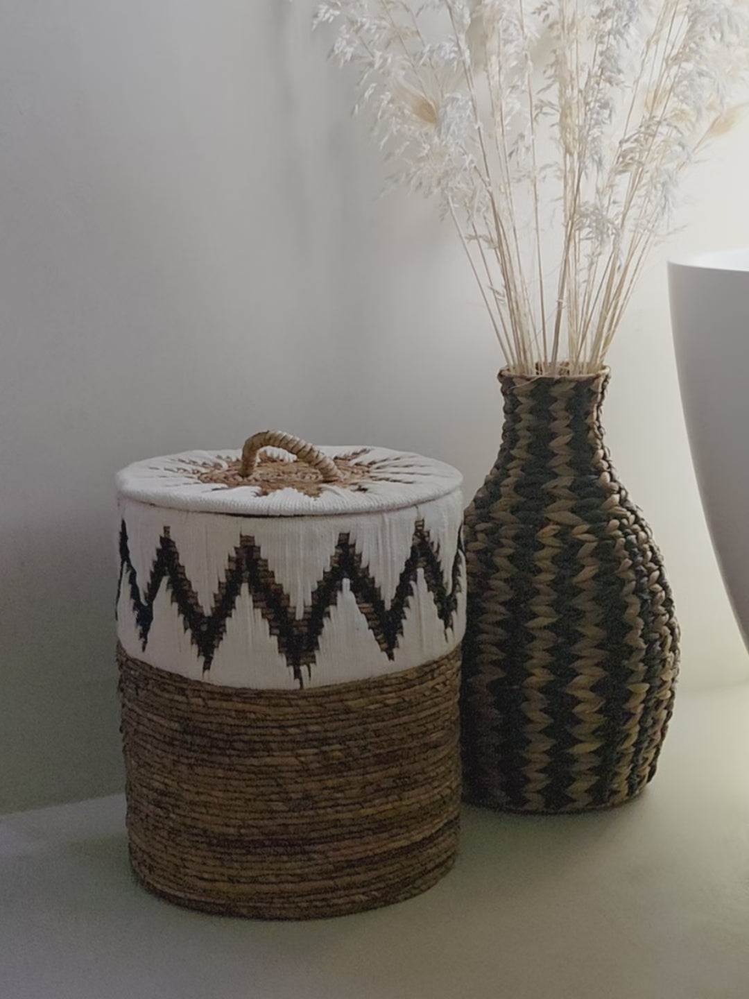 Samuel White handwoven abaca storage basket featuring a distinctive black and white zigzag pattern, a fitted lid and a decorative handle, styled beside the Zahara woven vase in a bright, natural bathroom interior.