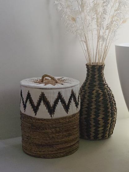 Samuel White handwoven abaca storage basket featuring a distinctive black and white zigzag pattern, a fitted lid and a decorative handle, styled beside the Zahara woven vase in a bright, natural bathroom interior.