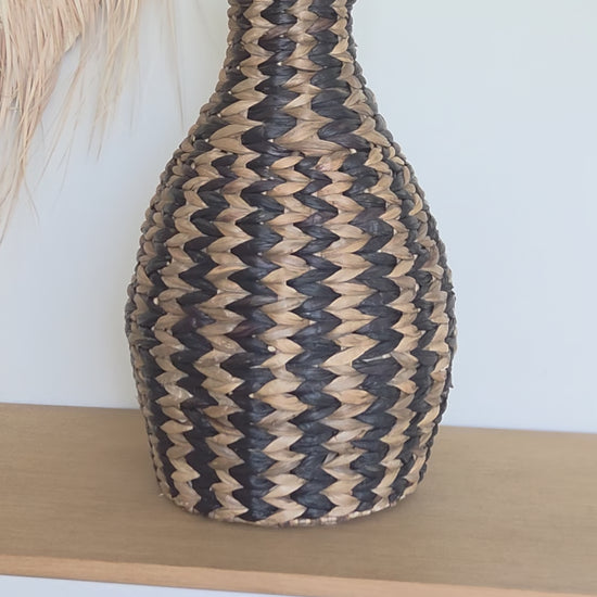 Close-up video of the Zahara decorative woven vase, highlighting its distinctive black and natural zigzag pattern, rounded silhouette and narrow neck. Styled with dried grasses on a wooden cabinet in a bright interior.