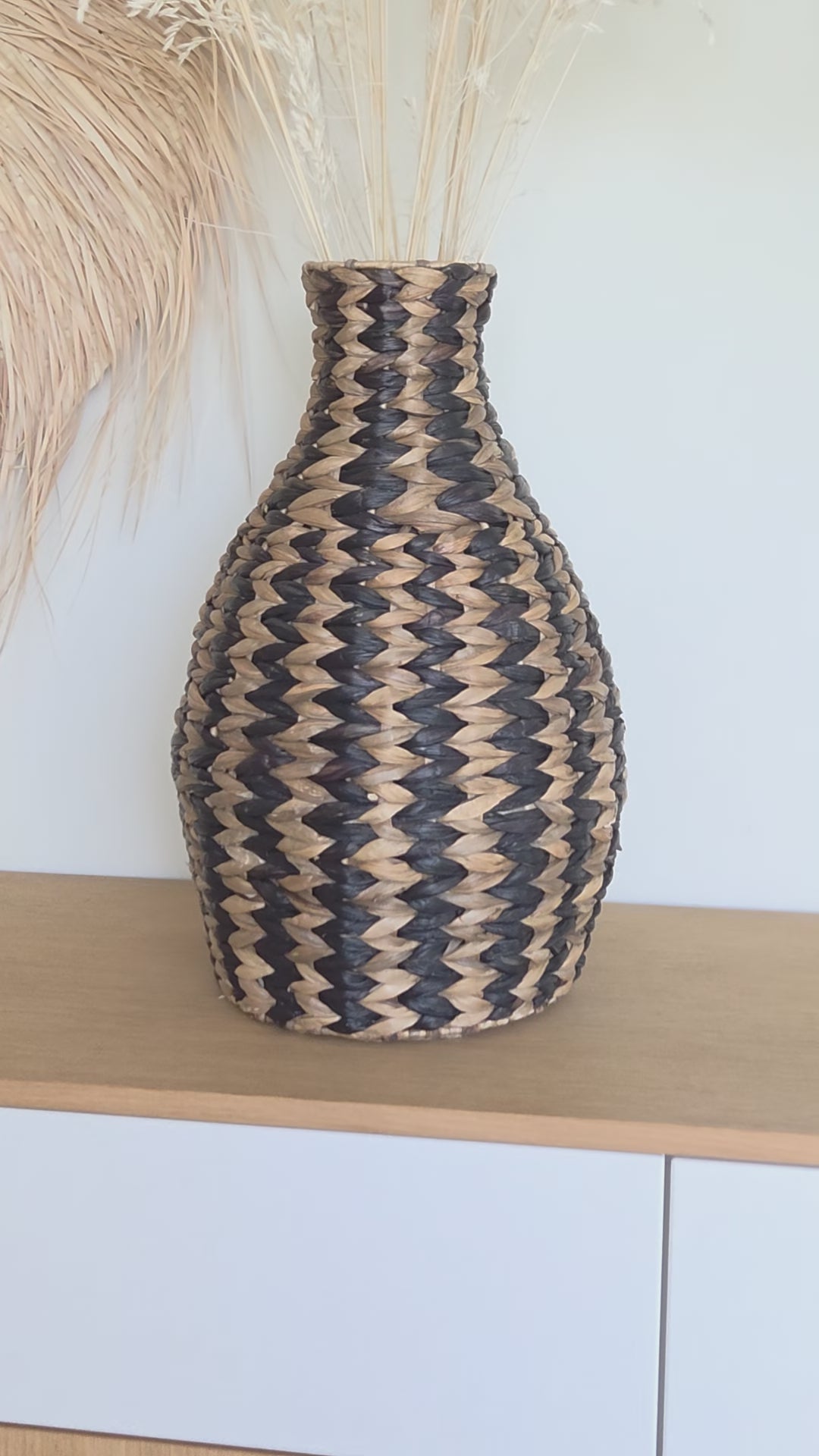 Close-up video of the Zahara decorative woven vase, highlighting its distinctive black and natural zigzag pattern, rounded silhouette and narrow neck. Styled with dried grasses on a wooden cabinet in a bright interior.