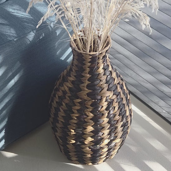 Zahara decorative woven vase featuring a bold black and natural zigzag pattern, filled with dried grasses and styled beside a dark sofa in a sunlit living room. The video highlights its distinctive shape and textured weave