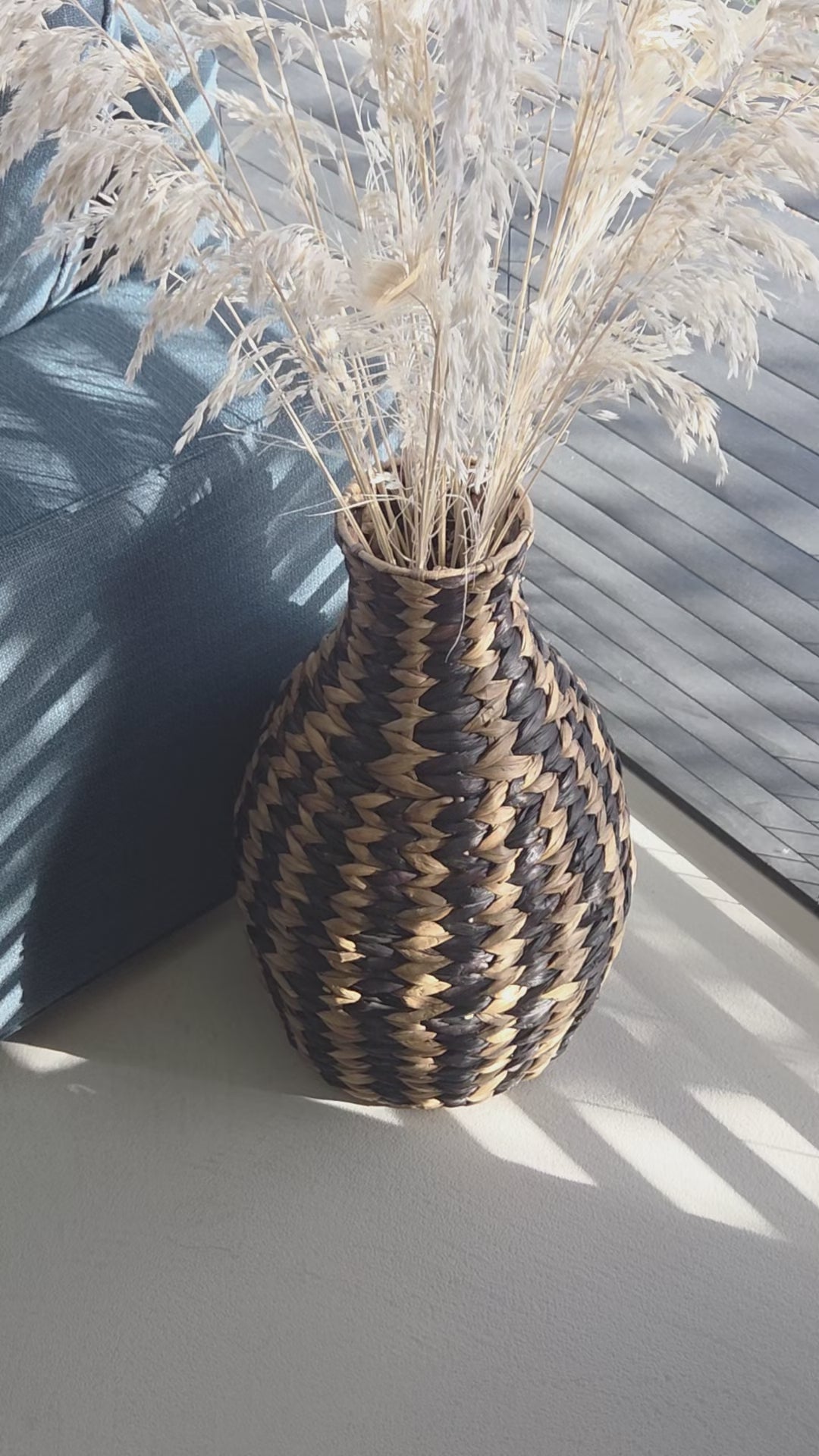 Zahara decorative woven vase featuring a bold black and natural zigzag pattern, filled with dried grasses and styled beside a dark sofa in a sunlit living room. The video highlights its distinctive shape and textured weave