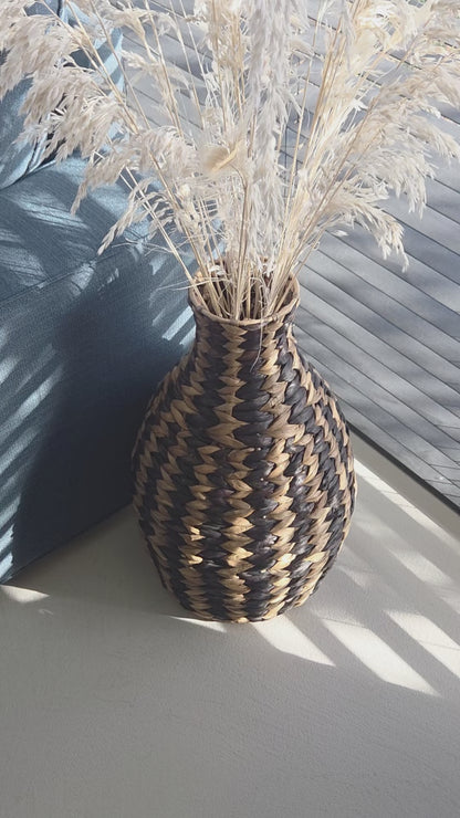 Zahara decorative woven vase featuring a bold black and natural zigzag pattern, filled with dried grasses and styled beside a dark sofa in a sunlit living room. The video highlights its distinctive shape and textured weave